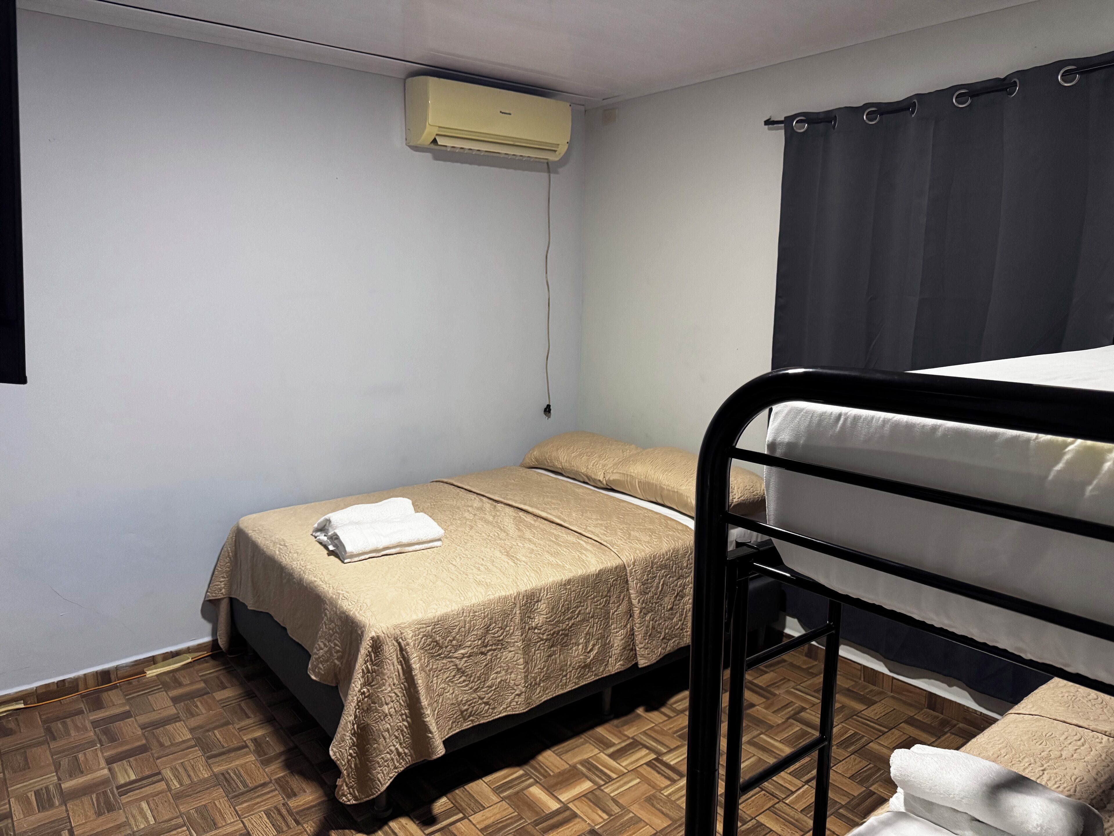 A small, single-bed room with a wooden floor, a bed with a beige bedspread, a black metal headboard, a window with a dark curtain, and an air conditioning unit on the wall.
