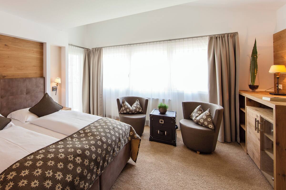 Une chambre d’hôtel avec un grand lit, deux fauteuils, une petite table avec une lampe, une table de chevet et une armoire en bois.
