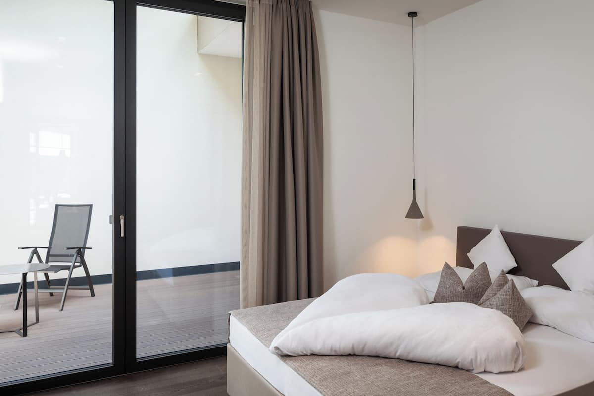 Une chambre d’hôtel moderne, équipée d’un lit, d’une chaise et offrant une vue sur l’extérieur grâce à une porte-fenêtre coulissante.