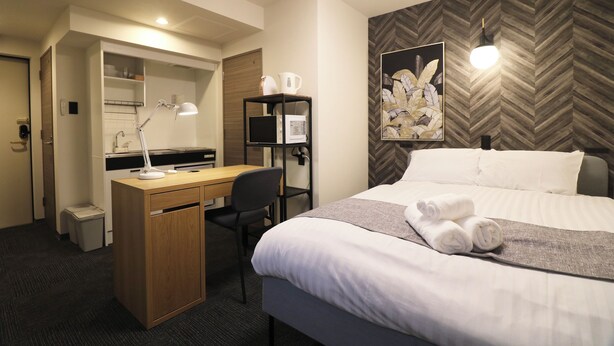 A hotel room with a bed, a desk with a lamp, a chair, a small trash bin, and a wall-mounted shelf with a kettle and cups. - TAKETO STAY PREMIERE 201 (Sapporo)