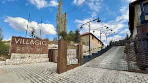 En skylt med texten ”VILLAGIO RESORT”, placerad vid en mur av sten och en stenlagd gångväg som leder fram till en byggnad.