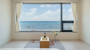 Een kamer met een groot raam met uitzicht op zee, een houten tafel met twee witte mokken, en een vaas met bloemen.