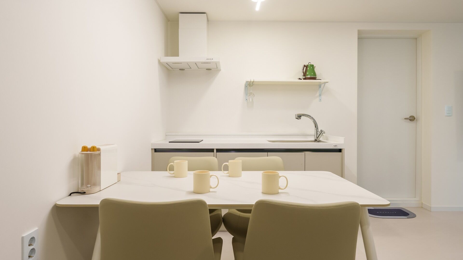 A small, clean kitchen with a white countertop, a sink, and a green kettle on a shelf. There is a table with four chairs and four mugs.