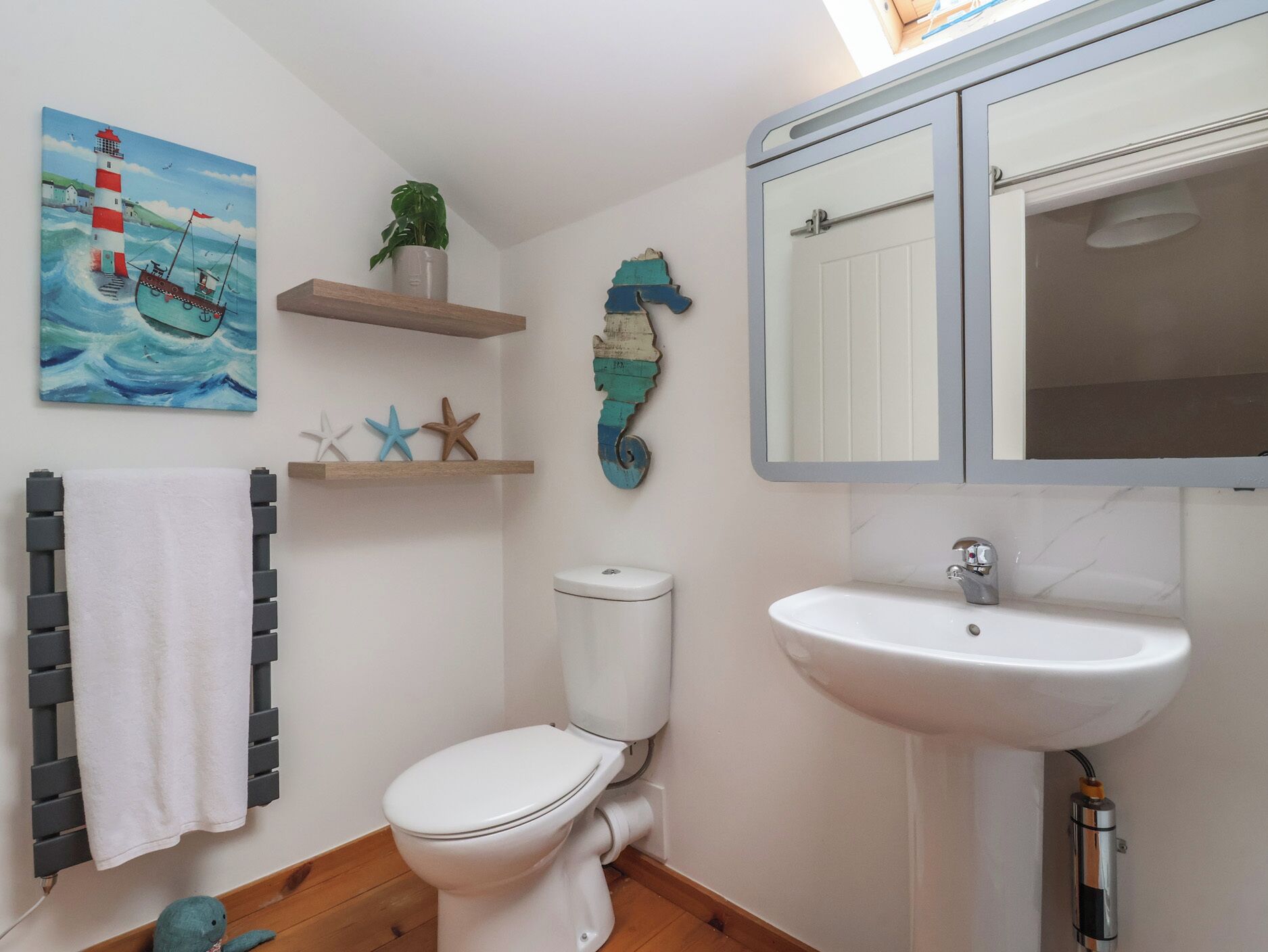 Een badkamer met een wit toilet, een witte wastafel, een spiegel, een handdoekhouder, een schilderij van een vuurtoren en een boot, en een klein plankje met een plant en zeesterren.