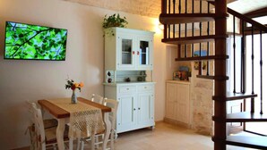 A dining area with a table and chairs, a white cabinet with glass doors, a wooden staircase, and a television mounted on the wall. - Arcaica cummersa - Dimora in pietra (Alberobello)