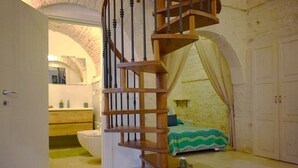 A bedroom with a wooden spiral staircase, a bed with a green and blue striped bedspread, a cactus, and a bathroom with a white toilet and sink. - Arcaica cummersa - Dimora in pietra (Alberobello)