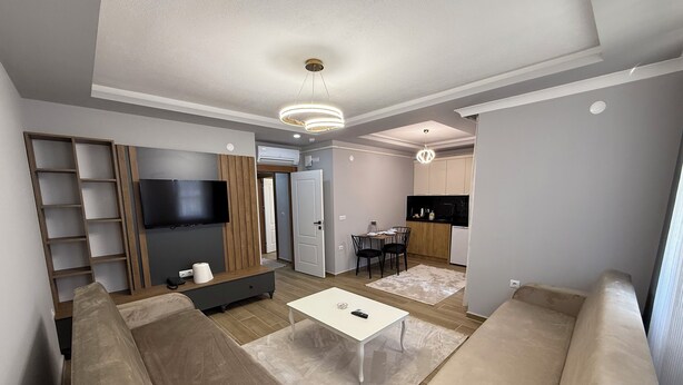 Een moderne woonkamer met een zithoek, een televisie, een houten accentmuur en een keuken in de achtergrond.