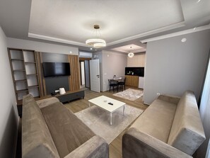 A modern living room with a sofa set, a TV, a wooden accent wall, and a kitchen area in the background. - Güven Palace Suites (Sürmene)
