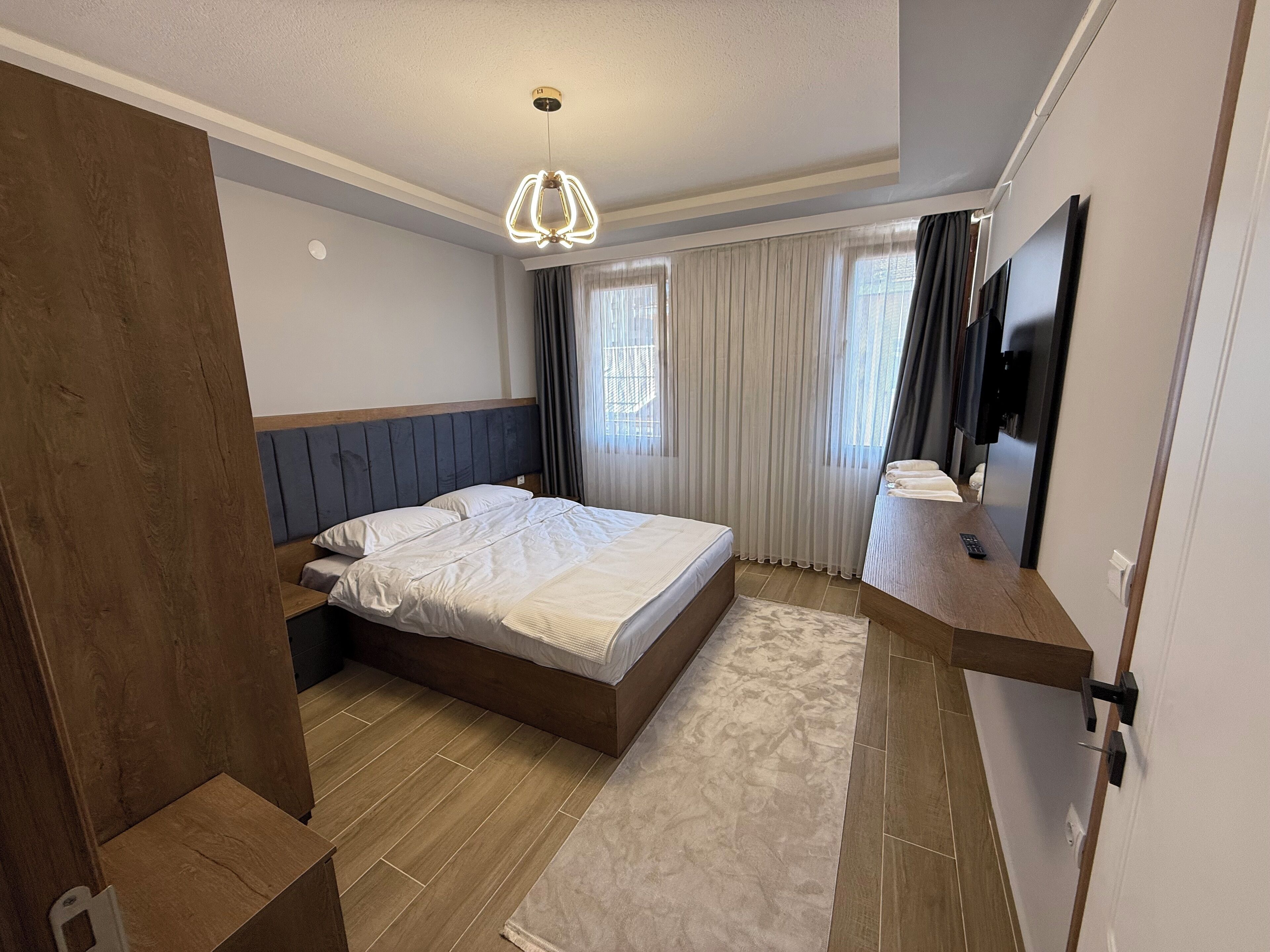 A modern hotel room with a large bed, a wooden headboard, a bedside table with a lamp, a large mirror, and a view of the city through the window.