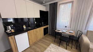 Een moderne keuken met een inbouwoven, een koffieapparaat en een kleine koelkast. Er is een zithoek met een tafel voor vier personen, een bank en een raam met gordijnen.