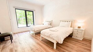 Un dormitorio con dos camas, una silla, una mesita de noche con lámpara y una ventana con vistas al verdor.