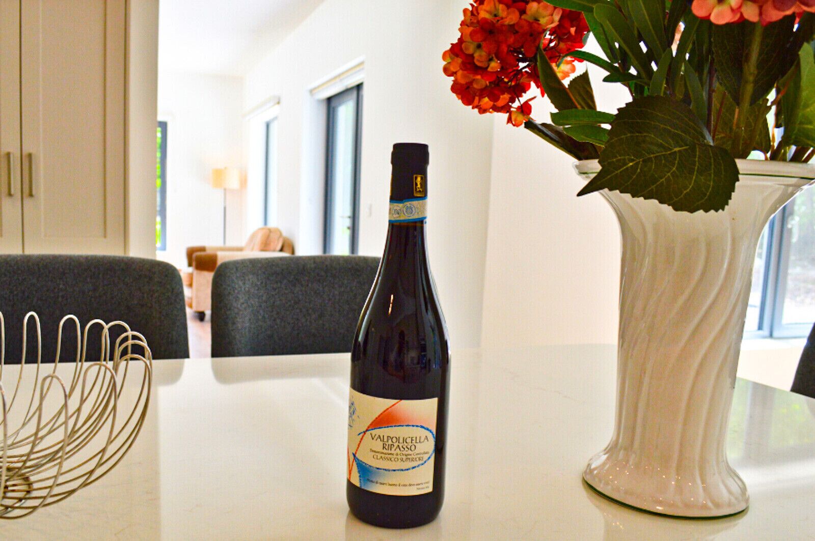 A bottle of Valpolicella Ripasso wine on a table with a vase of flowers and a wire basket.