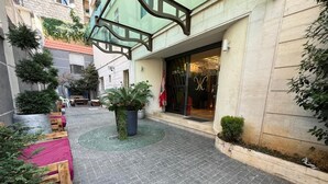 Une cour avec un sol carrelé, des bancs en bois, des plantes en pot et une entrée d’immeuble dotée d’une porte en verre.