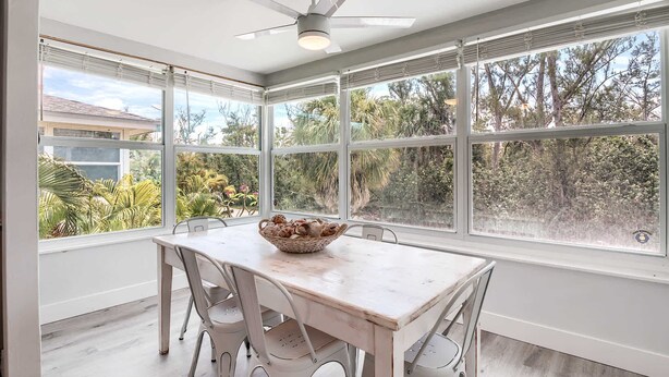 Un espace repas comprenant une table et des chaises, de grandes fenêtres donnant sur un paysage verdoyant et un ventilateur de plafond.