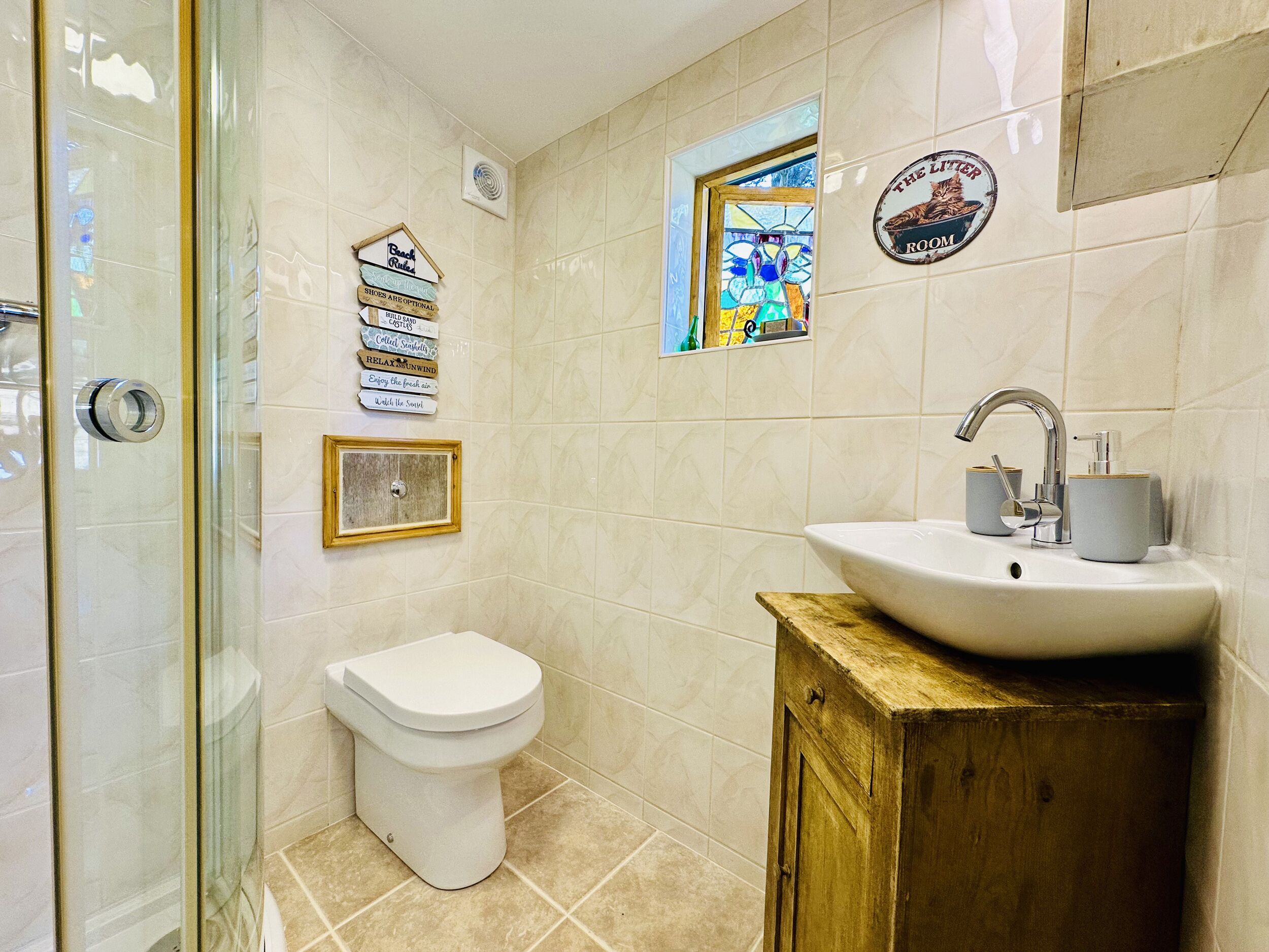 Ein Badezimmer mit Toilette, Waschbecken und Dusche. An der Wand befinden sich Dekorationen, darunter ein gerahmtes Bild und ein Schild mit verschiedenen Aufschriften.