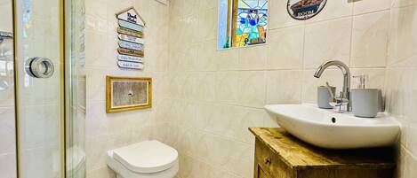 Ein Badezimmer mit Toilette, Waschbecken und Dusche. An der Wand befinden sich Dekorationen, darunter ein gerahmtes Bild und ein Schild mit verschiedenen Aufschriften.