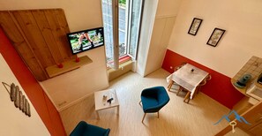 Ein kompakter Wohnraum mit Fernseher, einem Esstisch, zwei Stühlen, einem kleinen Tisch mit Fernbedienung und einem Fenster mit Blick auf die Straße.