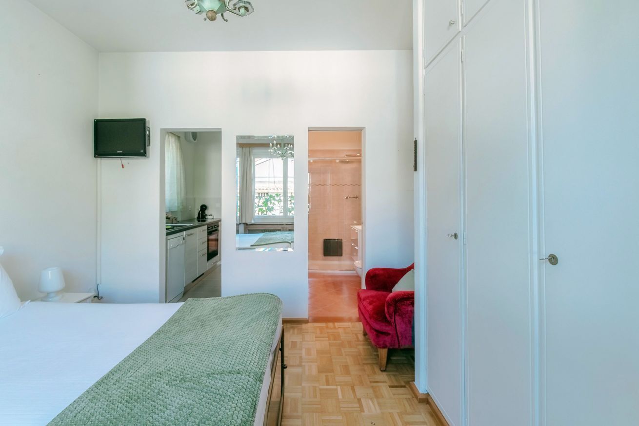 A bedroom with a bed, a TV, a red chair, and a view of a kitchen and bathroom through an open door.