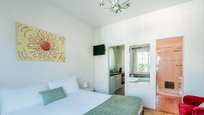 A bedroom with a bed, a green throw, a red chair, a TV, and a bathroom visible through an open door.