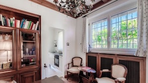 A dining area with a wooden table, two chairs, a chandelier, and a large window with a view of greenery.