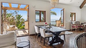 A spacious room with a wooden dining table, white chairs, a chandelier, and large windows offering a view of the ocean.