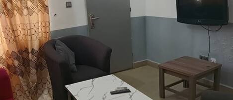 Une petite pièce simple, avec un lit, une chaise, une table, un téléviseur et une porte.