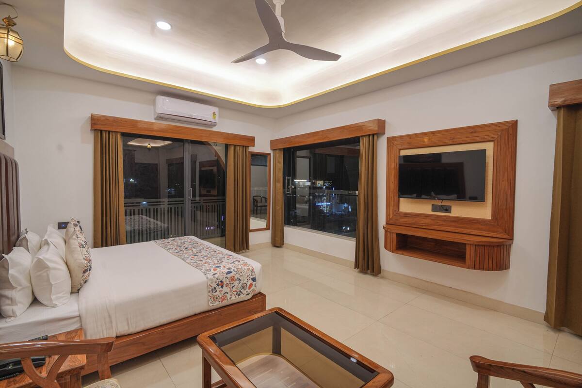 Habitación de hotel con cama, televisor montado en la pared, ventilador de techo y ventanas amplias.