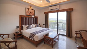 Une chambre d’hôtel équipée d’un lit, d’une table, d’une chaise, d’un climatiseur, d’un ventilateur de plafond, d’un tableau et d’un balcon avec vue.