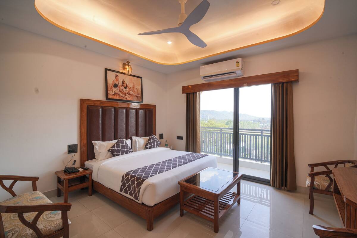Habitación de hotel con cama, mesa, silla, equipo de aire acondicionado, ventilador de techo, un cuadro y un balcón con vistas.