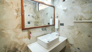 Une salle de bain avec un mur en marbre, un lavabo rectangulaire, un miroir avec un cadre en bois et deux appliques murales.