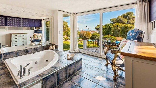 A bathroom with a large bathtub, a shower, and a sink. There is a window with a view of a garden and a balcony with a table and chairs. - Luxury 5-Bedroom Villa Gardens Heated Pool, Jacuzzi & Sea Views – Estepona (Bahía Dorada)