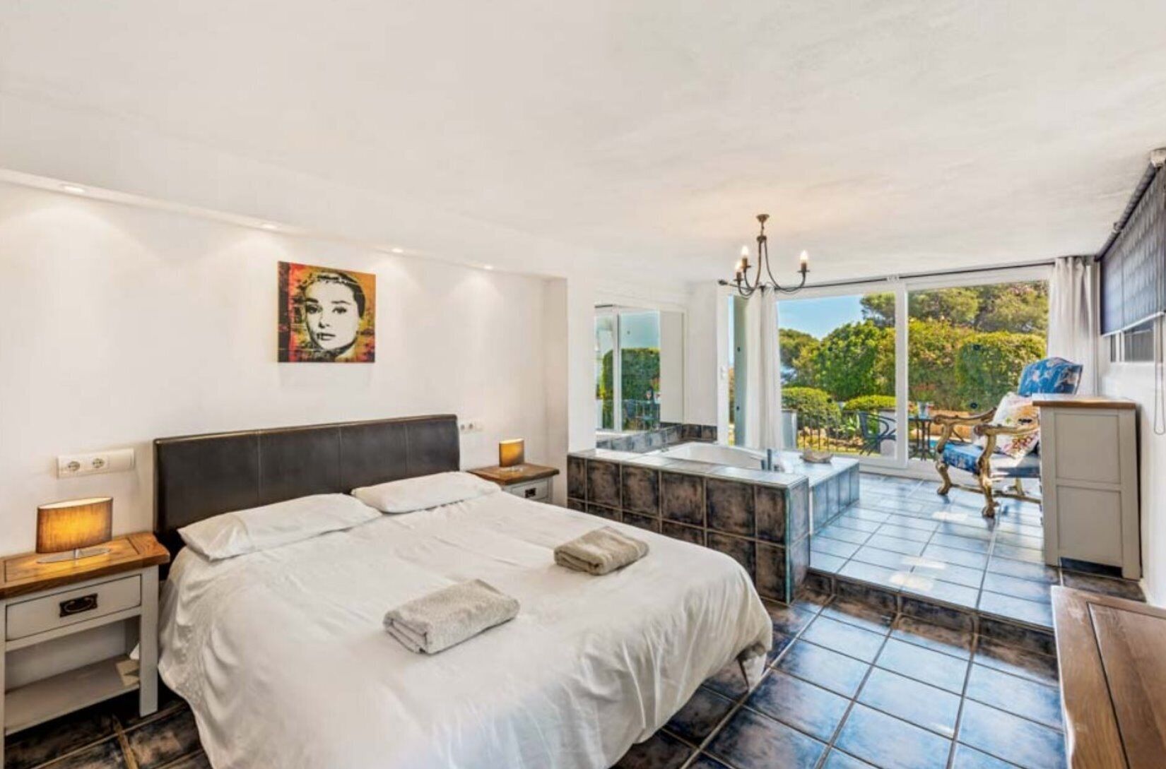 A bedroom with a large bed, a bathroom with a bathtub, and a balcony with a view of greenery.