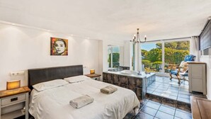 A bedroom with a large bed, a bathroom with a bathtub, and a balcony with a view of greenery. - Luxury 5-Bedroom Villa Gardens Heated Pool, Jacuzzi & Sea Views – Estepona (Bahía Dorada)