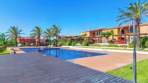 A swimming pool with a wooden deck, surrounded by palm trees and a building with a red roof.
