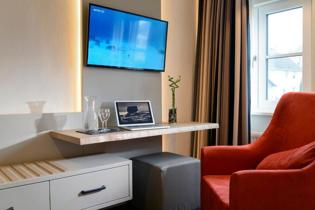 Hotelkamer met een wandgemonteerde flatscreen televisie, een laptop op een houten plank, een rode fauteuil, een klein nachtkastje met een glas en een raam met gordijnen.