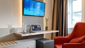 Ein Hotelzimmer mit einem am Wand montierten Flachbildfernseher, einem Laptop auf einem Holzregal, einem roten Sessel, einem kleinen Nachttisch mit einem Glas und einem Vorhangfenster.