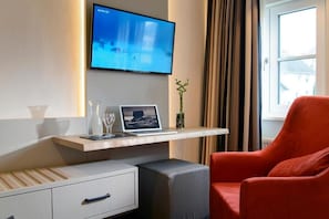 A hotel room with a flat-screen TV mounted on the wall, a laptop on a wooden shelf, a red armchair, a small bedside table with a glass, and a window with curtains. - Landgasthof Kleiner  (Sundern)
