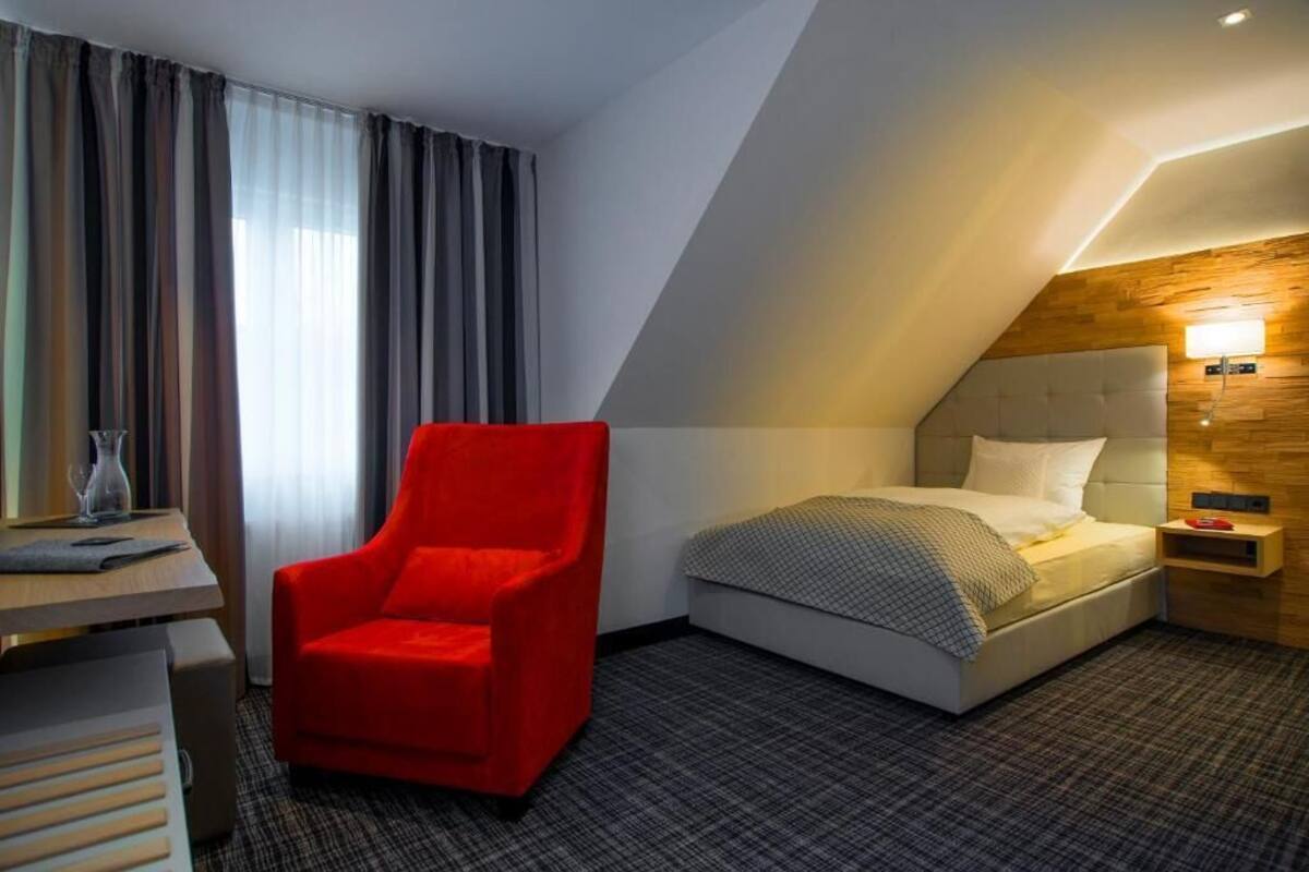 Una habitación de hotel con una cama, un sillón rojo, un escritorio y una ventana con cortinas.