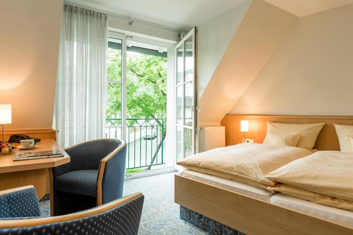 Habitación de hotel con cama, escritorio con silla, ventana con cortinas y balcón con vista a árboles.