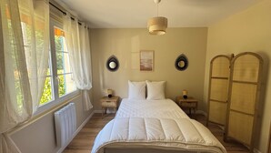 A bedroom with a large bed, two bedside tables with lamps, a ceiling light, and a view of greenery through the window.