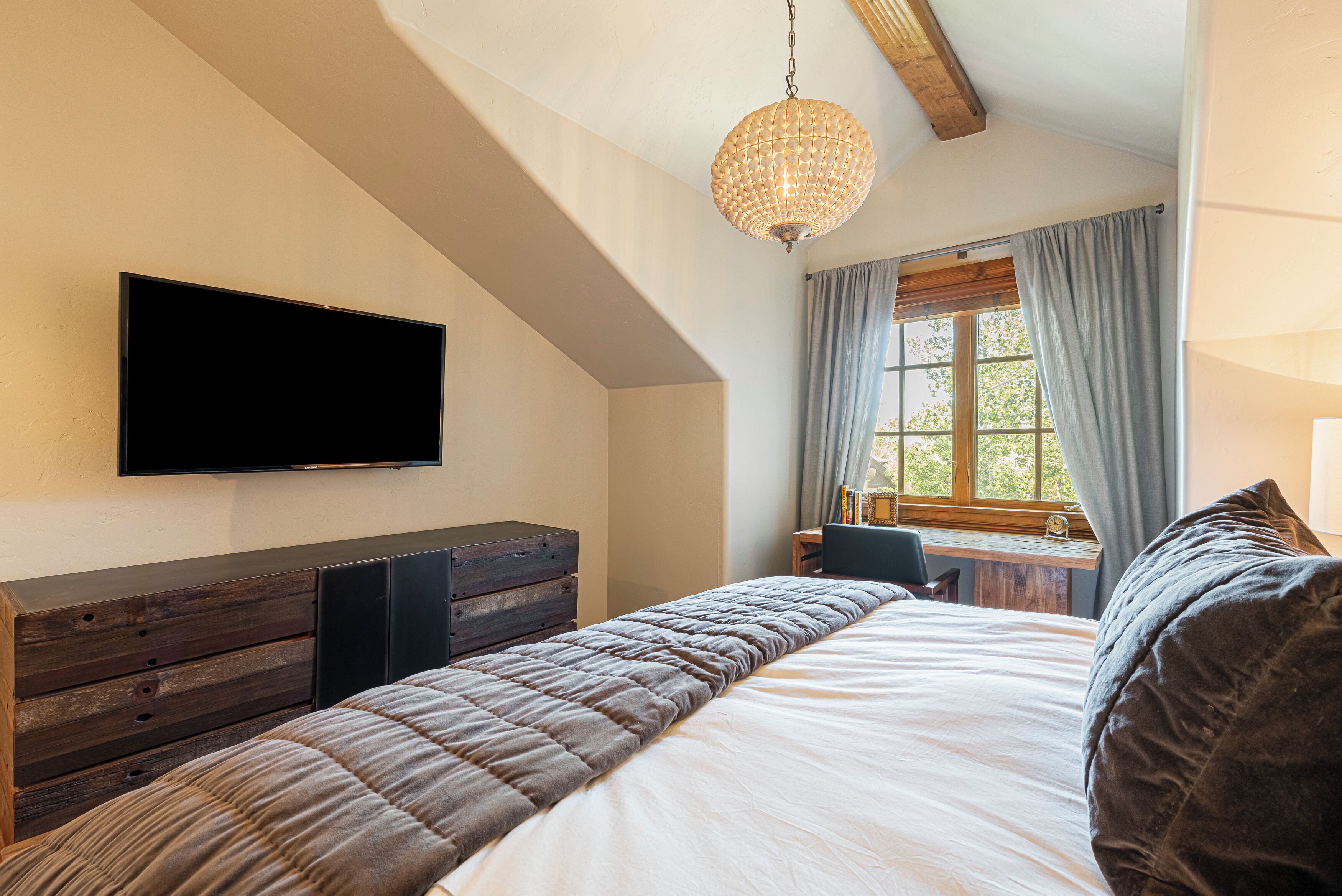 Une chambre à coucher avec un grand lit, un téléviseur à écran plat fixé au mur, une commode en bois et une fenêtre avec des rideaux.