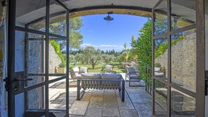 A covered outdoor seating area with stone flooring, a bench, and a view of a garden with trees and a stone wall. - Mas Cicada (Paradou)
