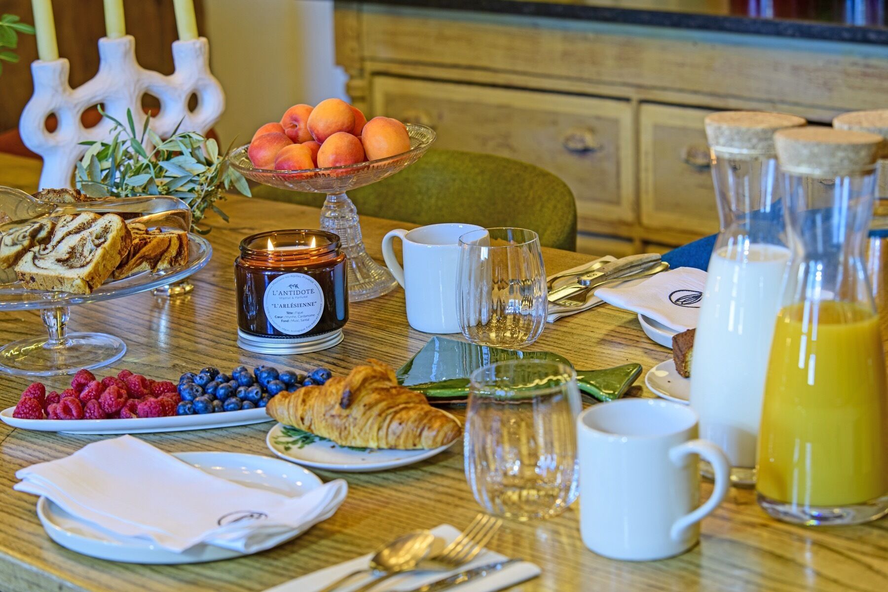 Une table de petit-déjeuner avec des viennoiseries, des fruits, une bougie et un verre de jus d’orange.