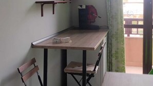 Ett litet rum med ett skrivbord i trä, två stolar, en tv som är monterad på väggen och ett fönster med gardiner.