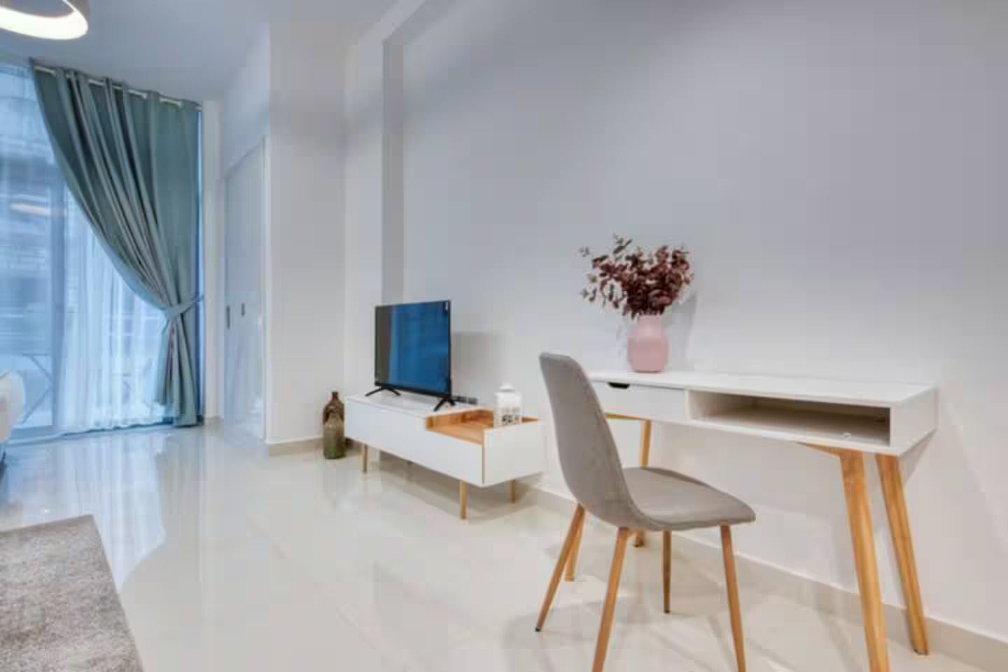A modern living room with a flat-screen TV, a white console table, a grey chair, and a vase with dried flowers.