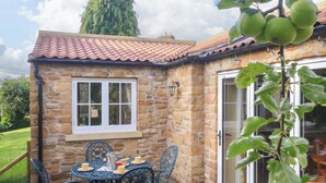 A stone building with a tiled roof, a window, and a door. There is a table with chairs and a plant with green leaves. - Apple Tree Cottage (York)