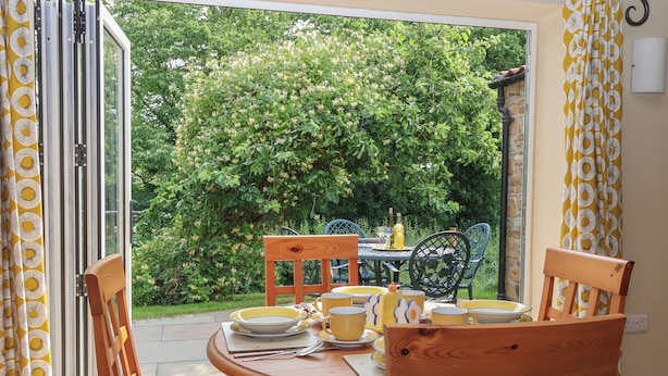 A dining area with a table set for two, chairs, and a view of a garden with trees and a stone wall. - Apple Tree Cottage (York)