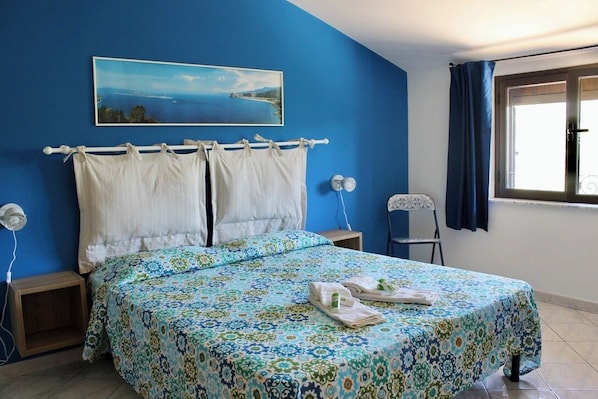 A bedroom with a bed, a nightstand, a chair, a window with curtains, and a painting on the wall.