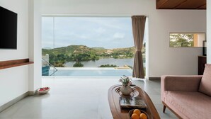 Un salon moderne doté d’une grande fenêtre donnant sur un lac et des collines, d’un téléviseur à écran plat fixé au mur et d’un coin salon confortable.