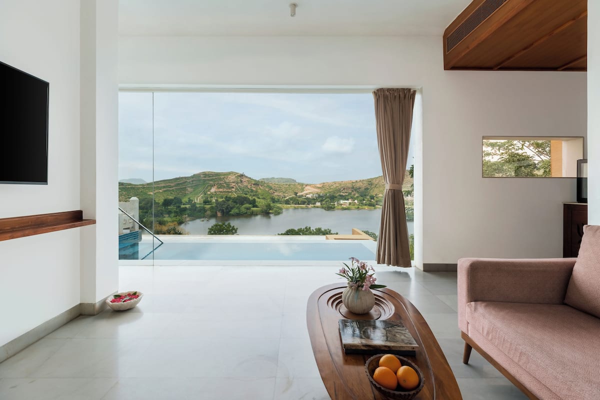 Una sala de estar moderna con un amplio ventanal con vistas a un lago y colinas, un televisor de pantalla plana montado en la pared y una zona de estar acogedora.